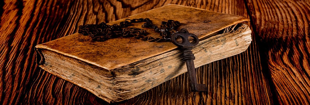 An old and damaged book, with an iron chain and key sits on a heavy wood grain table.